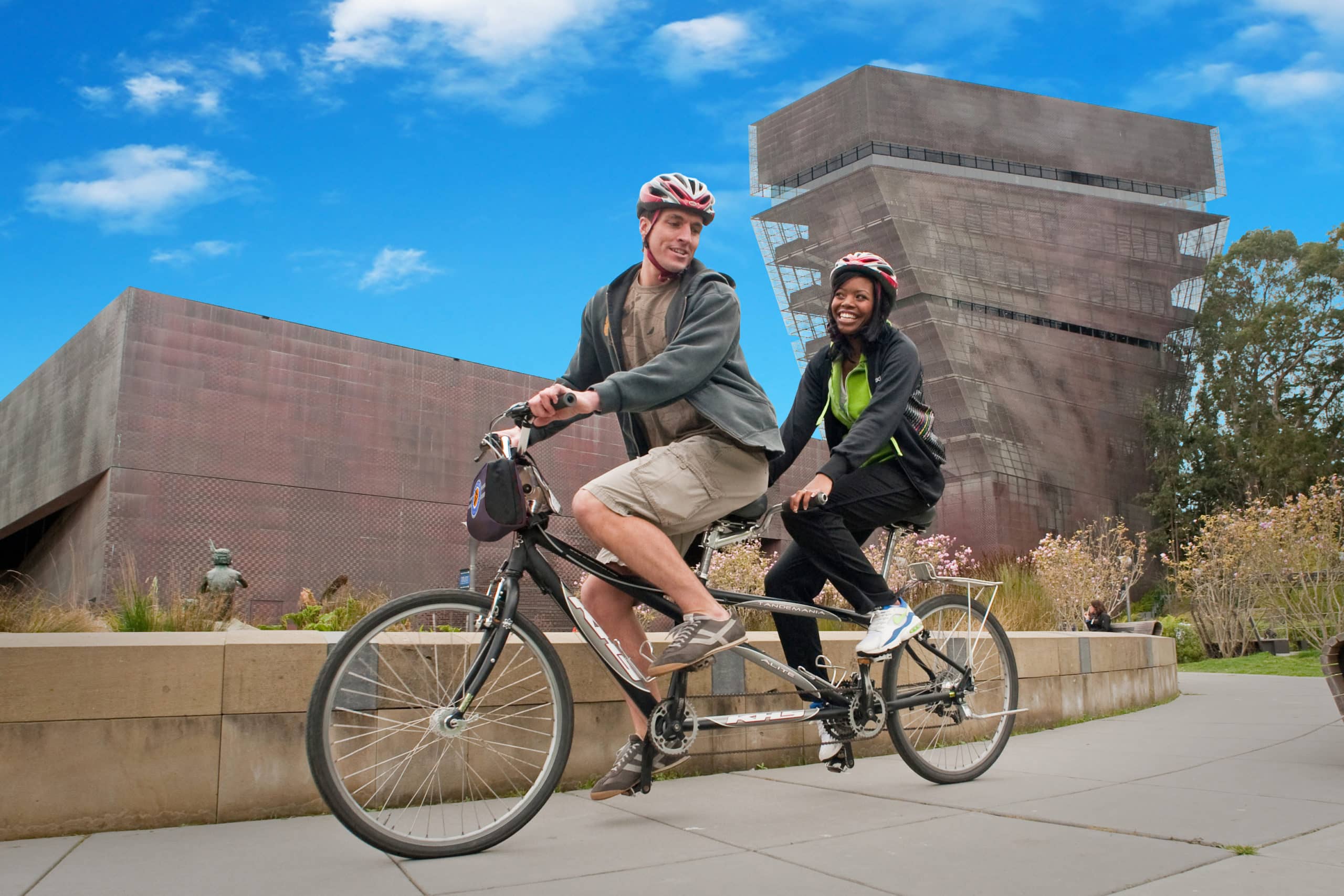 two people on a tandem bike pass in front of sf's de young musem
