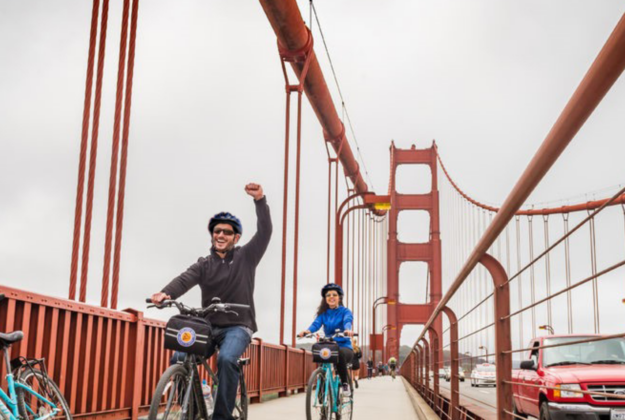 A man holds up his arm in victory as he and another rider bike across the Golden Gate Bridge