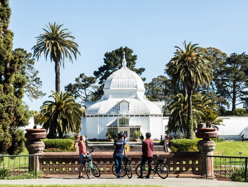Three bikers hold their bikes and stand in front a white building in Golden Gate Park