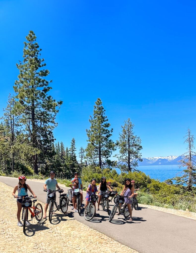 A group of people hold their bike rentals and smile in front of beautiful pine trees near Lake tahoe